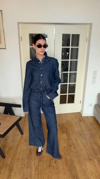 Sötétkék farmer overált viselő személy napszemüvegben és sportcipőben, bent állva egy fehér üvegezett dupla ajtó mellett, fa padlón.