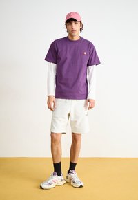 Camiseta de manga corta morada superpuesta sobre una camisa de manga larga blanca, combinada con pantalones cortos gris claro y zapatillas multicolores. Gorra rosa.