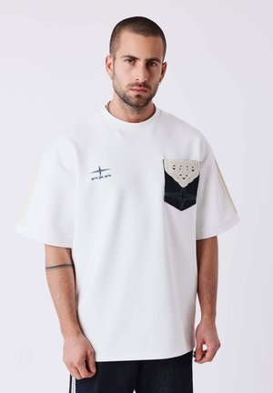 Homme aux cheveux courts et foncés avec une barbe, portant un T-shirt blanc oversize avec une poche noire et un détail en crochet beige.