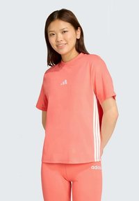 Kortärmad idrottströja i korallfärg, med vit Adidas-logotyp och tre vita ränder på varje sida. Tillverkad av mjukt och andningsbart material.