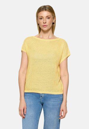 Kvinde med glat blondt hår iført en kortærmet gul striktop og blå jeans, stående mod en ensfarvet lys baggrund.