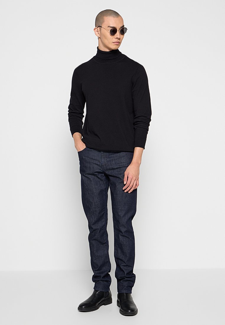 Schwarzer Rollkragenpullover, dunkelblaue Jeans und schwarze Knöchelstiefel. Das Model trägt runde Sonnenbrillen und steht mit den Händen in den Taschen.