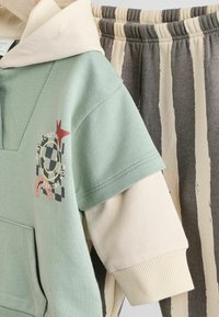 Mintgrüne Kapuzenjacke mit einer cremefarbenen Kapuze und grafischen Details, kombiniert mit leichten grauen und cremefarbenen senkrecht gestreiften Hosen. Weicher Stofftextur.