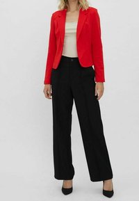 Blazer rouge cintré porté sur un haut beige, associé à un pantalon noir taille haute à jambes larges. Des talons à bout pointu complètent la tenue.