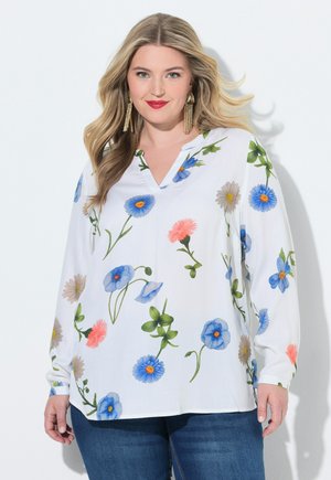 Kobieta z długimi blond włosami, nosząca złote kolczyki z frędzlami i białą bluzkę z niebiesko-różowym kwiatowym wzorem, połączoną z niebieskimi dżinsami.