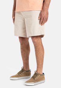 Pantalones cortos de pana beige con un corte recto, que llegan a la mitad del muslo. Combinados con zapatos de ante marrón que tienen suelas de goma blanca y cordones.