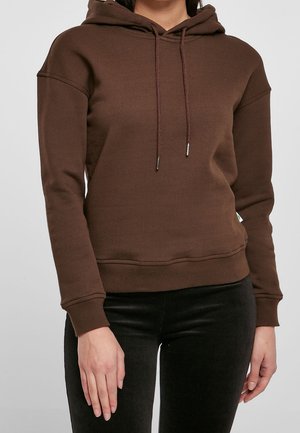Personne portant un sweat à capuche marron uni à manches longues avec des cordons, associé à un pantalon noir, debout devant un fond uni.