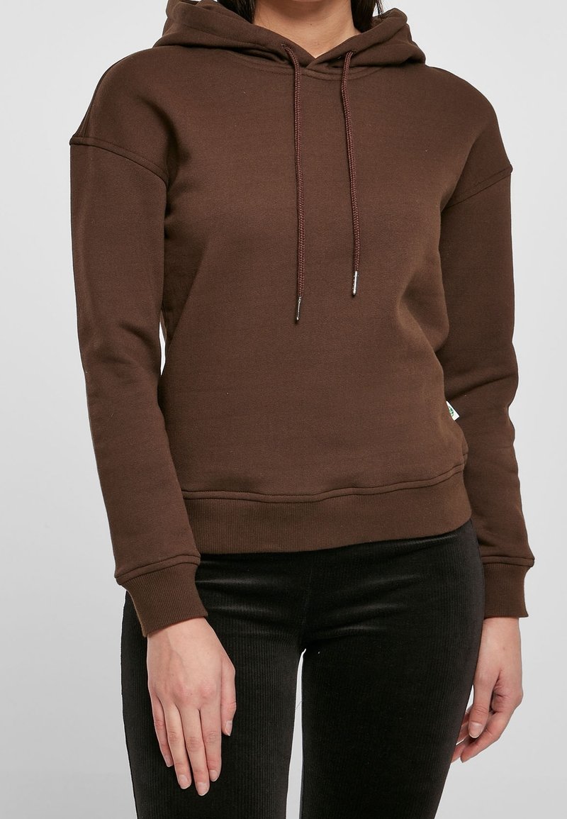 Personne portant un sweat à capuche marron uni à manches longues avec des cordons, associé à un pantalon noir, debout devant un fond uni.