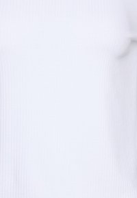 Ribbad vit topp gjord av ett slätt, stretcht tyg, med en figurnära silhuett. Texturen är fin med subtila vertikala linjer.