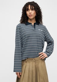 Vrouw met krullend haar, gekleed in een marineblauw-wit gestreept poloshirt met lange mouwen en een beige geplooide rok, staand met één hand op haar heup.