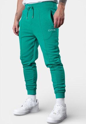 Mann trägt türkisfarbene Jogginghosen mit Kordelzug in der Taille und Reißverschlusstaschen an den Seiten, kombiniert mit weißen Sneakers und Socken, stehend vor weißem Hintergrund.