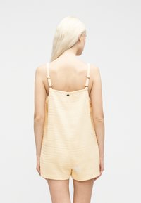 Γυναίκα  με μακριά ξανθά μαλλιά, φοράει ένα υφασμάτινο, αμάνικο ολόσωμο σορτς σε ανοιχτό μπεζ χρώμα, στέκεται με την πλάτη προς τα εμπρός σε απλό λευκό φόντο.