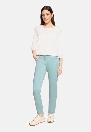 Femme debout avec une main dans la poche d’un pantalon bleu pastel, portant un haut blanc manches longues et des baskets beiges sur un fond uni.