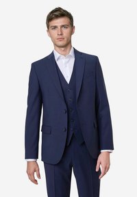 Ein junger Mann steht selbstbewusst da, trägt einen maßgeschneiderten marineblauen Anzug mit einem weißen Hemd und zeigt einen gelassenen Gesichtsausdruck vor einem schlichten Hintergrund.