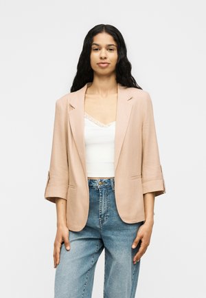 Mlada žena s dugom crnom kosom nosi bež sako preko bijelog čipkastog topa i plave traperice visokog struka, stoji na bijeloj pozadini.