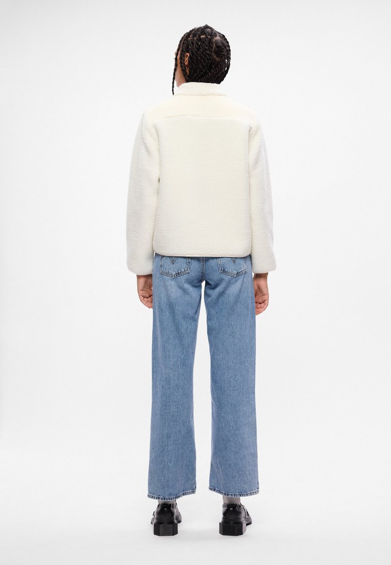 Hvid fleecejakke med en struktureret overflade, kontrasterende glatte skuldre, parret med lyseblå brede jeans og sorte platformstøvler.