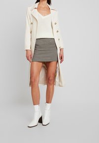 Kvinde iført en cremefarvet trenchcoat over en hvid strikbluse og en sort-hvid houndstooth mini-nederdel med hvide hælede ankelstøvler.