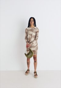 Prilegla obleka z dolgimi rokavi v rjavem tie-dye vzorcu, v kombinaciji z zeleno polmesecasto torbo, ki ima napis "Juicy", in platform sandali.