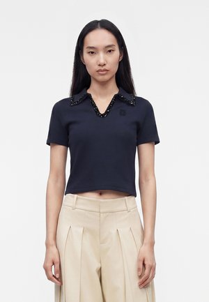 Kvinne iført en marineblå kortermet topp med perlebesatt krage og beige høytlivsplisserte bukser mot en ensfarget hvit bakgrunn.