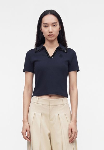 Femme portant un haut à manches courtes bleu marine avec un col orné de perles et un pantalon beige plissé taille haute sur un fond blanc uni.