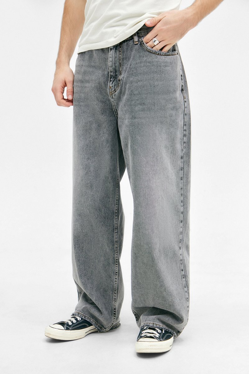 Person iført løse grå jeans, sort og hvide sneakers, hvid t-shirt, med én hånd i lommen og en sølvring på fingeren.