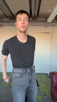 Bărbat tânăr purtând un tricou negru și blugi gri, stă în interior lângă o ușă albă și un scaun de piele roșie, pe o mochetă cu model verde.