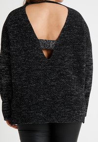 Žena nosící černý a šedý mramorovaný pletený svetr s hlubokým výstřihem do V na zádech a odpovídajícím pásem přes horní část zad.