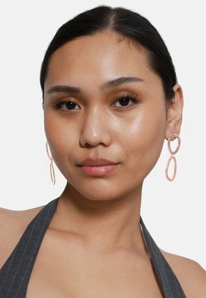 Boucles d'oreilles créoles en or rose avec des formes ovales superposées. Fini lisse, conception légère et style minimaliste. Le modèle porte un haut à col halter.