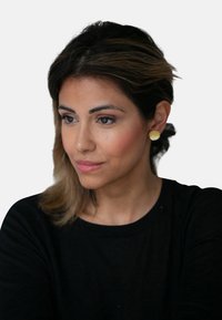 Gulltonede ovale øreringer med en glatt tekstur. Modellen har på seg en svart skjorte, med lange mørke hår samlet bak, som viser frem øreringene.