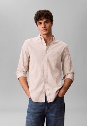 Νέος άντρας με καστανά μαλλιά φοράει ανοιχτόχρωμο ριγέ πουκάμισο με μακριά μανίκια που έχουν γυριστεί και μπλε τζιν, στέκεται με τα χέρια στις τσέπες.