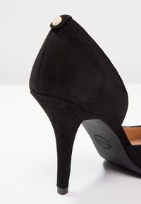 Chaussure à talon haut en daim noir avec talon pointu, détail bouton doré près de l'arrière, et semelle lisse avec logo de la marque.