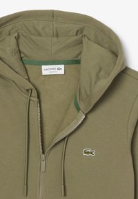 Felpa con zip color verde oliva realizzata in tessuto morbido, con cappuccio foderato, dettagli verdi e logo del coccodrillo Lacoste sul petto.