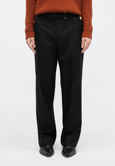 Pantalons larges noirs à la coupe ajustée, dotés d'une passante de ceinture, d'une texture lisse et de plis repassés à l'avant, associés à des chaussures noires.