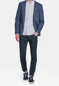 Marinblå texturerad kavaj över en ljusgrå skjorta med knappar, parat med mörka jeans och marinblå sneakers med grå detaljer och rullade ärmar.