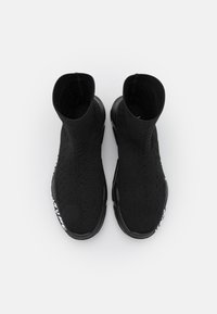 Zapatos de estilo calcetín negros con parte superior de punto texturizado, forma curvada y suelas de goma negras con letras blancas de la marca en el lateral.