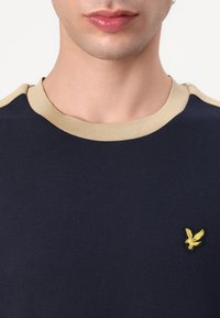 Tamno plava majica s okruglim izrezom i bež rebrastim detaljima. Sadrži mali žuti orao logotip na lijevoj strani prsnog koša. Materijal: pamuk.