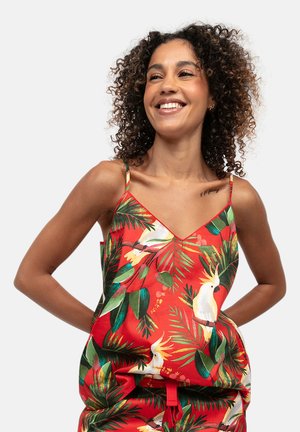 Donna sorridente con capelli ricci che indossa un top rosso senza maniche con foglie tropicali verdi e stampe di uccelli bianchi.