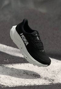 Chaussure de sport noire avec une tige texturée, logo blanc et semelle épaisse. Dispose d'un bout arrondi et d'un design épuré pour la course.
