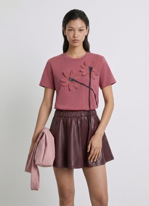 Donna con una t-shirt malva con applicazioni floreali e una gonna in pelle marrone scuro, che tiene in mano una giacca rosa piegata.