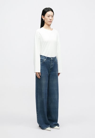 Biały top z długim rękawem w połączeniu z luźnymi, niebieskimi dżinsami z wysokim stanem. Modelka nosi białe, spiczaste szpilki. Prosta, czysta estetyka.