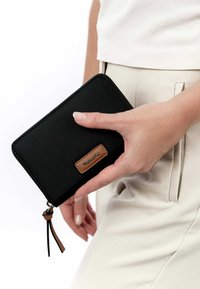 Schwarze Reißverschluss-Geldbörse mit strukturiertem Lederakzent und einem kleinen Lederzugband, in einer Hand gehalten, mit einem schlanken rechteckigen Design.