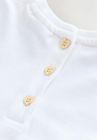 Hvid bomuldsbluse med tre gyldne hjerteformede knapper med indgraverede detaljer, placeret ved halsudskæringen med en glat tekstur.