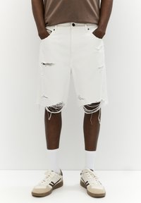 White denim shorts with frayed hems and distressing on the front, paired with a brown tee and white socks, worn with beige sneakers.