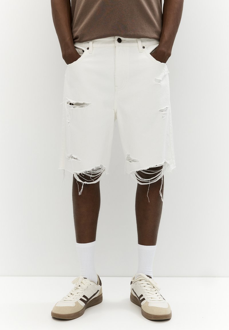 White denim shorts with frayed hems and distressing on the front, paired with a brown tee and white socks, worn with beige sneakers.