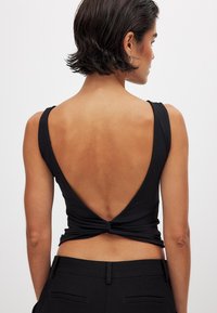 Schwarzes ärmelloses Oberteil mit tiefem V-Ausschnitt im Rücken, mit gerafftem Stoff an der Taille und glatter Textur. Kombination mit taillierten schwarzen Hosen.