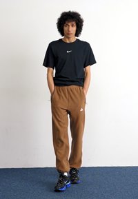 Black cotton t-shirt with white Nike logo, brown fleece joggers with side pockets, and black sneakers with blue accents, standing on a blue carpet.