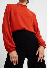 Blouse rouge à manches longues avec un col montant froncé et des poignets plissés, associée à un pantalon noir taille haute. Texture de tissu lisse.