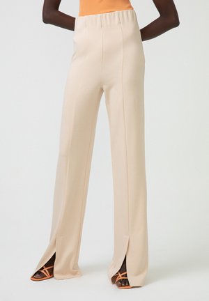 Light beige wide-leg pants with a stretchy waistband, side slits at the hem, made from a smooth material. Paired with strappy orange sandals.