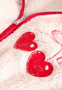 Jakna iz svetlo rožnate flis tkanine z rdečim zadrgo, z dvema rdečima vzorcema srca in belimi šivi kot poudarki. Mehka tekstura.