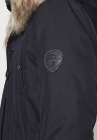 Marinblå parkasjacka med konstpäls, har knappstängning fram, dubbla bröstfickor och en rund logotypapplikation på ärmen. Slät textur.